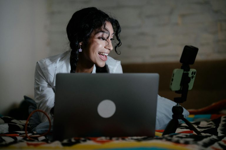 Happy woman vlogging with a laptop and smartphone setup, creating content indoors.